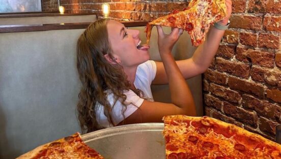 This photo shows a young woman with long brown hair and a white T-shirt holding an oversized pepperoni pizza slice above her head with both hands, ready to take a bite.