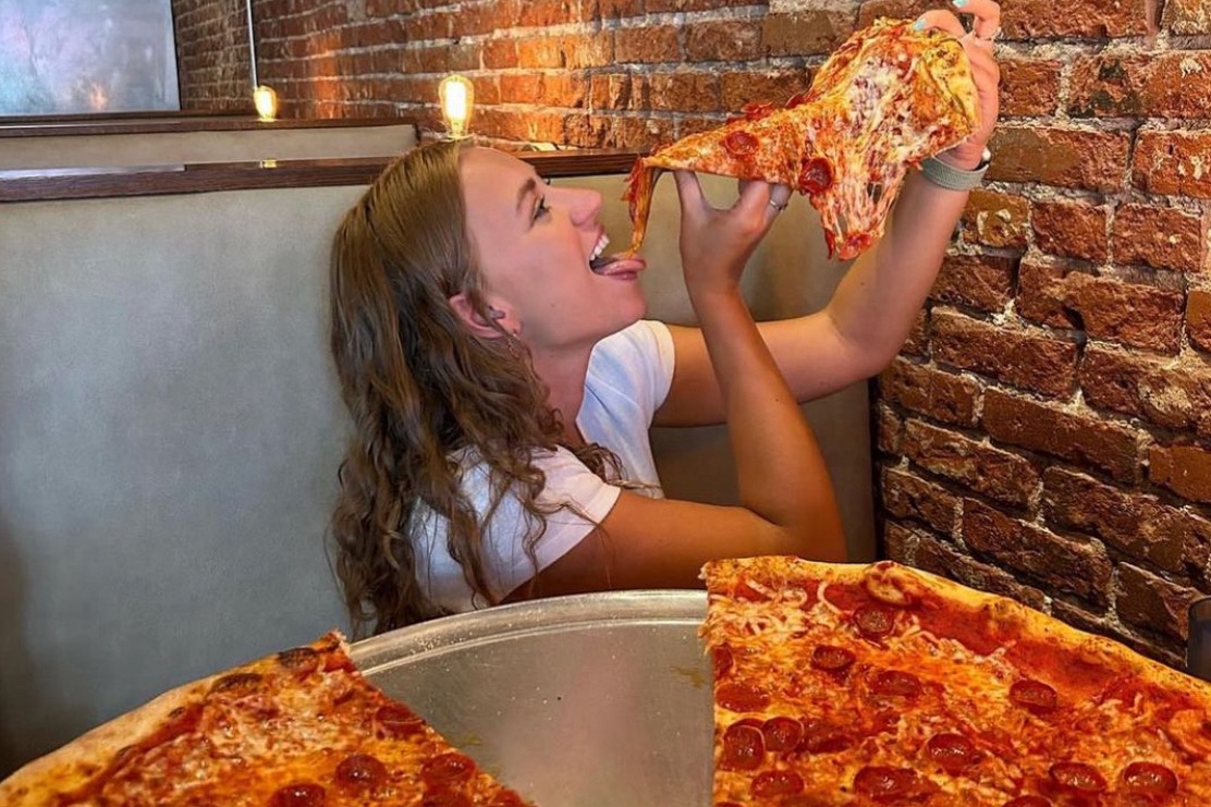 This photo shows a young woman with long brown hair and a white T-shirt holding an oversized pepperoni pizza slice above her head with both hands, ready to take a bite.