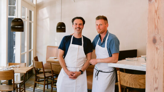 This photo shows Ryan Pollnow and Thomas McNaughton, both wearing long white aprons, standing together in the Flour & Water dining room, with McNaughton's arm around Pollnow.