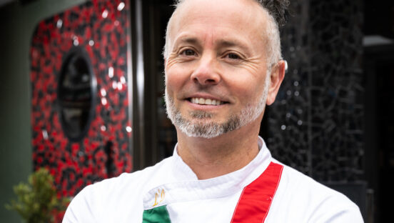 This photo shows Tony Gemignani smiling in a white chef coat with green and red stripes.