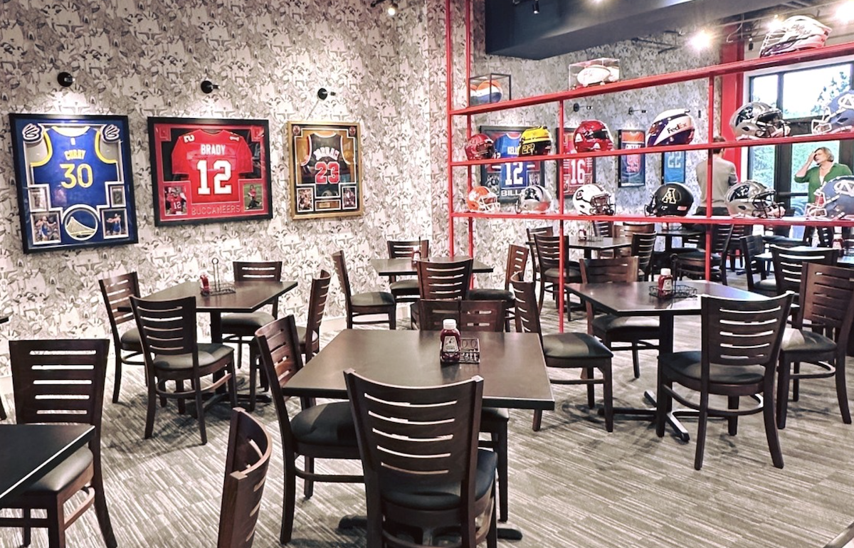 A new restaurant interior featuring sports memorabilia on the walls, including jerseys from athletes like Stephen Curry, Tom Brady, and Michael Jordan. There are also football helmets resting on a shelf.