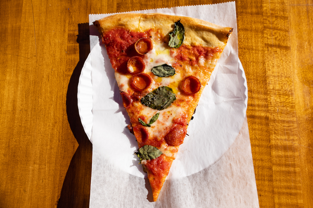 An overhead view of a New York City style pepperoni pizza slice topped with fresh basil on a white paper plate