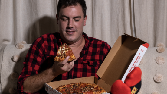 A man sits on a couch enjoying a slice of pizza while the pizza box sits in his lap.