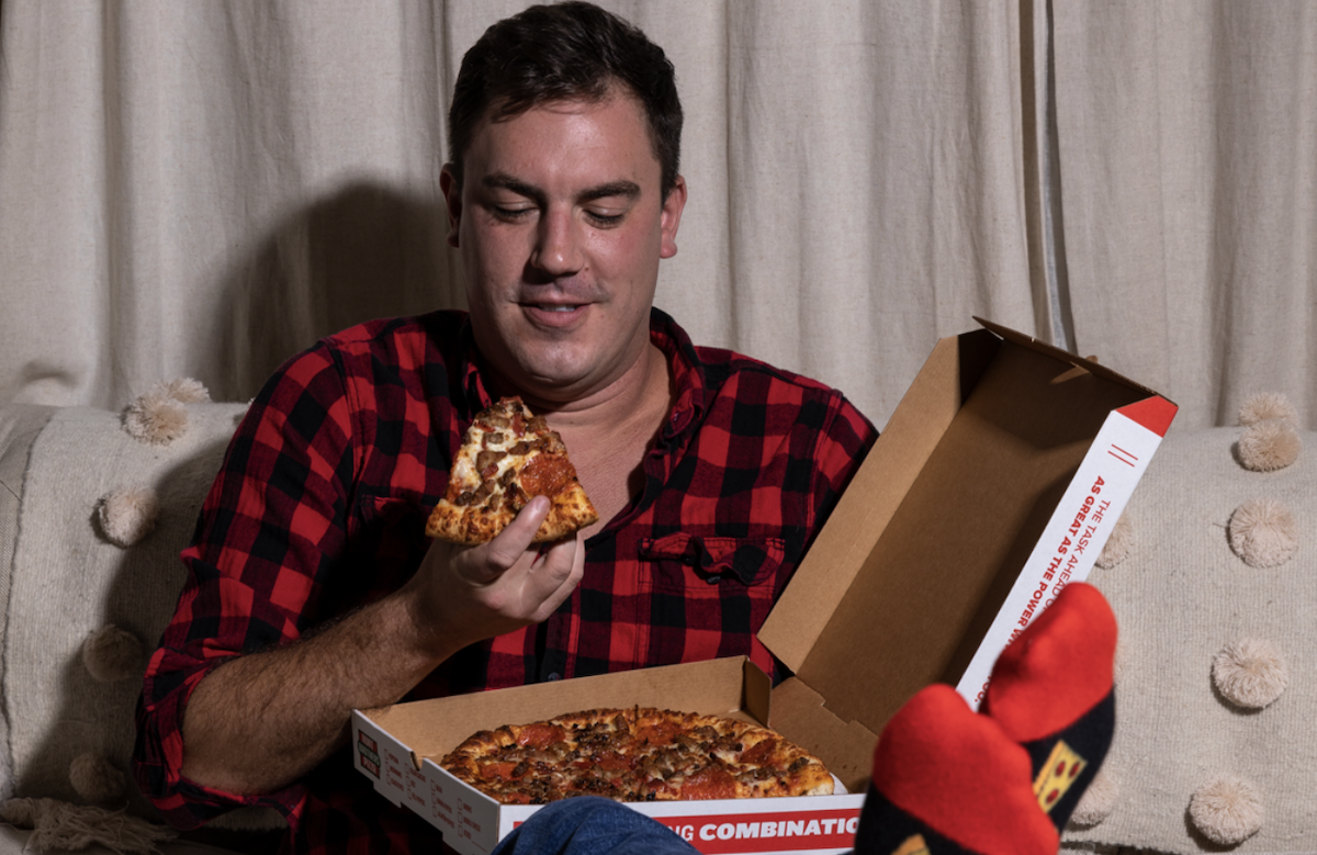 A man sits on a couch enjoying a slice of pizza while the pizza box sits in his lap.