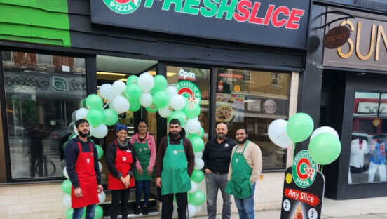 This photo shows a group of Freshslice employees standing together and holding a huge bouquet of green and white balloons in front of a new location.