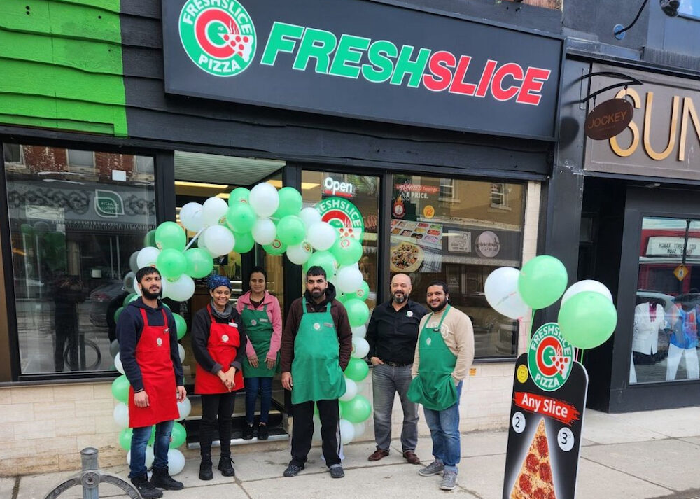 This photo shows a group of Freshslice employees standing together and holding a huge bouquet of green and white balloons in front of a new location.