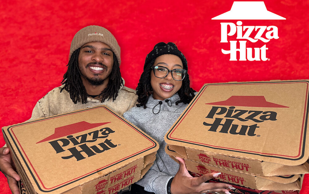 This photo shows Keith and Ronni Lee smiling and holding Pizza Hut logged boxes against a red background.