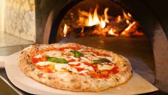 This photo shows a beautiful Neapolitan-style Margherita pizza on a wooden peel in front of a blazing wood-fired oven.