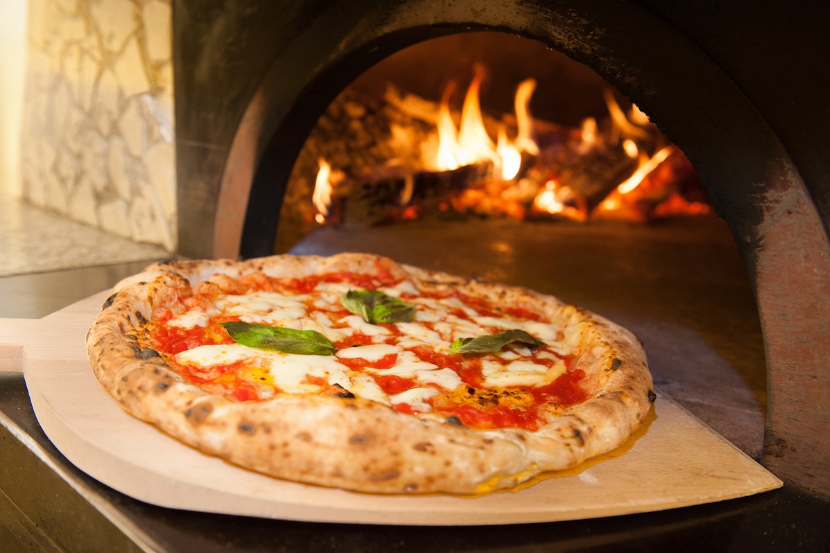 This photo shows a beautiful Neapolitan-style Margherita pizza on a wooden peel in front of a blazing wood-fired oven.