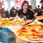This photo shows a young female MOD Pizza employee in a black T-shirt and red bandana, with several pizzas in front of her.