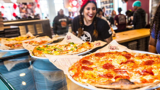 This photo shows a young female MOD Pizza employee in a black T-shirt and red bandana, with several pizzas in front of her.