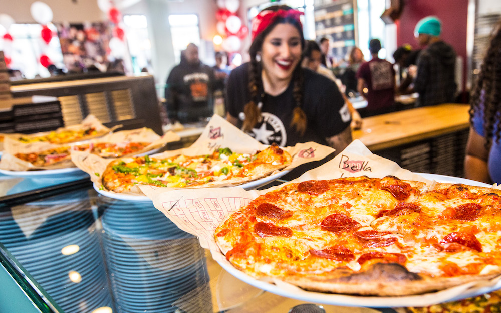 This photo shows a young female MOD Pizza employee in a black T-shirt and red bandana, with several pizzas in front of her.