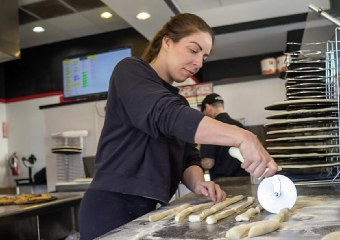 A pizzeria owner uses a pizza cutter to slash through dough in the kitchen of her shop.