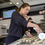 A pizzeria owner uses a pizza cutter to slash through dough in the kitchen of her shop.