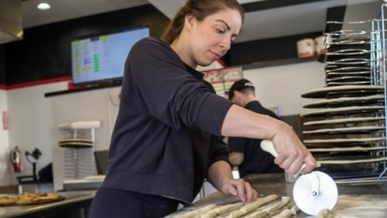 A pizzeria owner uses a pizza cutter to slash through dough in the kitchen of her shop.