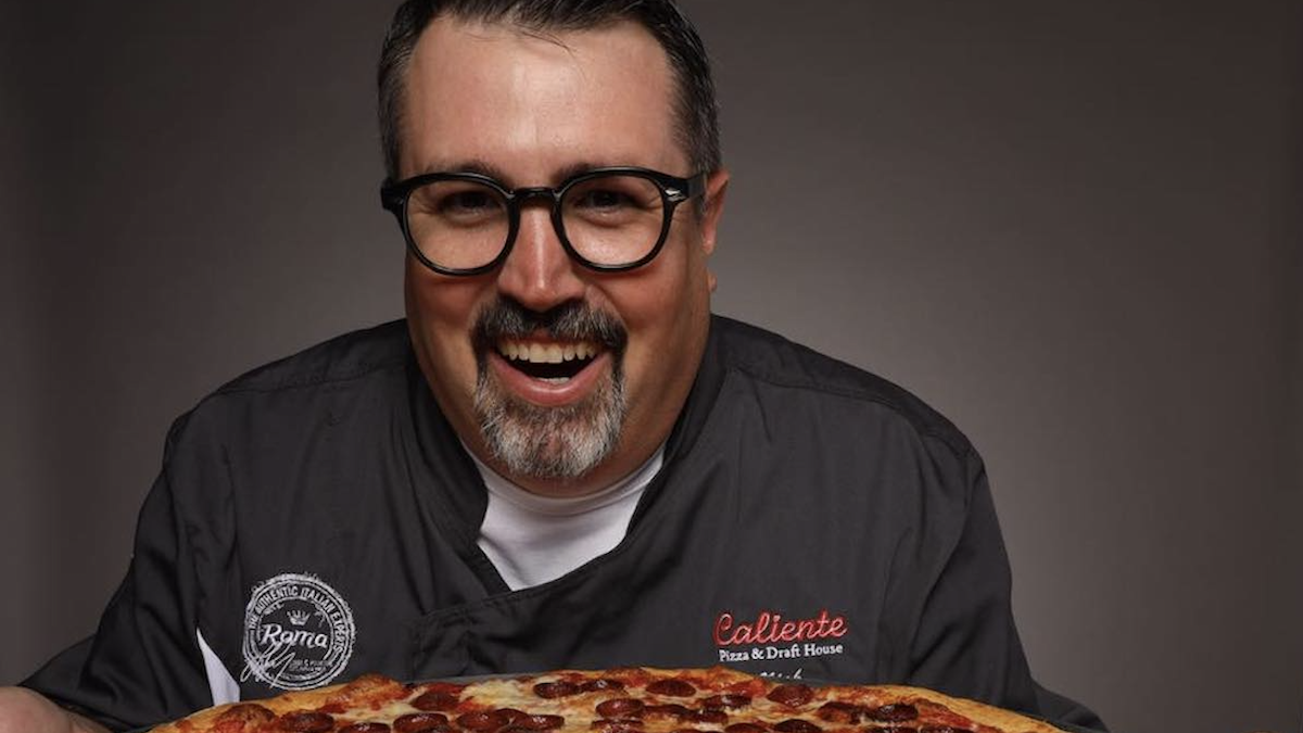 Nick Bogacz, owner of Caliente Pizza & Draft House, smiles as he looks at a pepperoni pizza in the foreground.
