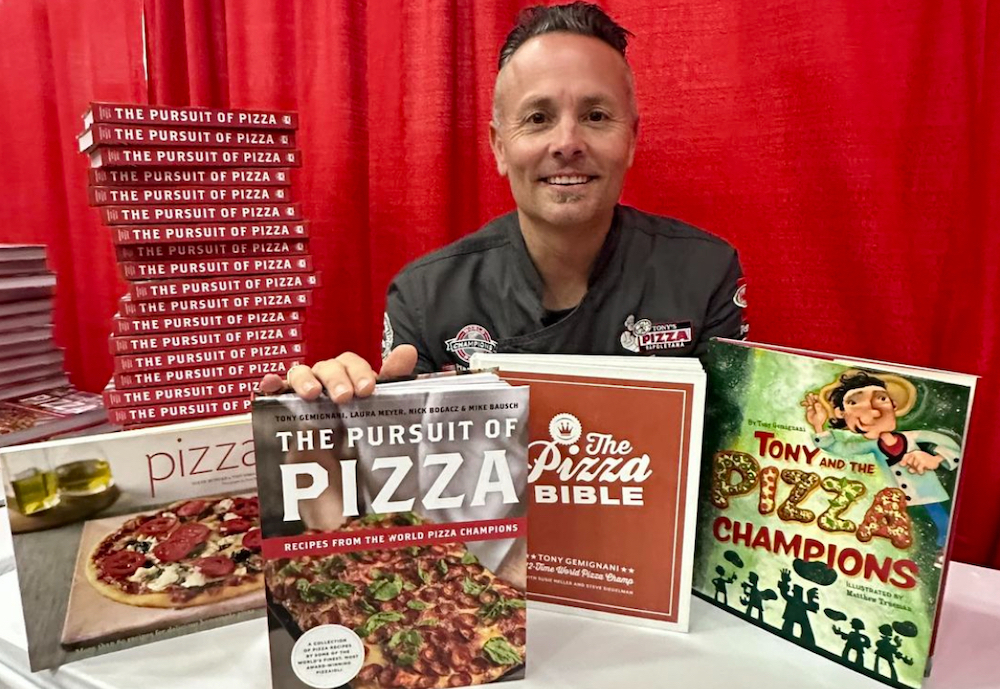 This photo shows Tony Gemignani in a black chef coat, sitting at a table with copies of his new cookbook and his older one.