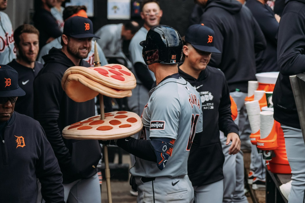 A photo of a Detroit Tigers baseball player standing in the Tigers' dugout holding a toy spear that is protruding through three fake pepperoni pizzas.