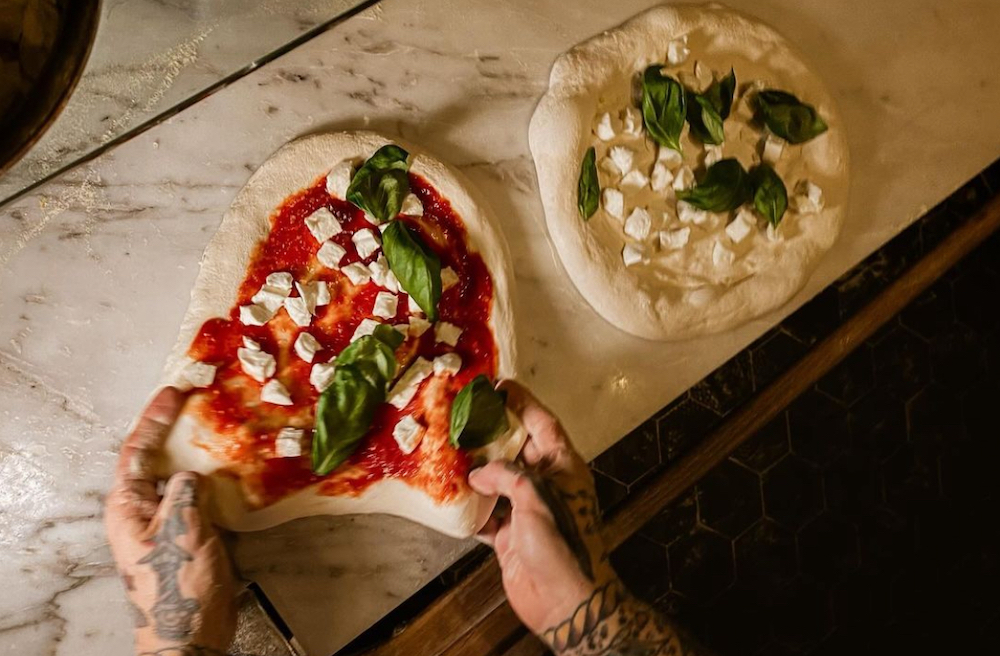 This photo shows a man's heavily tattooed hands and arms stretching out the dough for an unbaked pizza topped with sauce, cheese and basil. Next to it is a white-sauce pizza with cheese and basil as well.