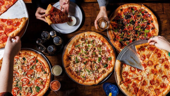 this photo shows a wide selection of pizzas arranged on a table at Village Idiot Pizza, with customers' hands shown pulling out slices.