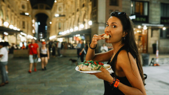 This photo shows a young, dark-haired Italian woman biting into a slice of pizza while walking on a busy street in Milan.