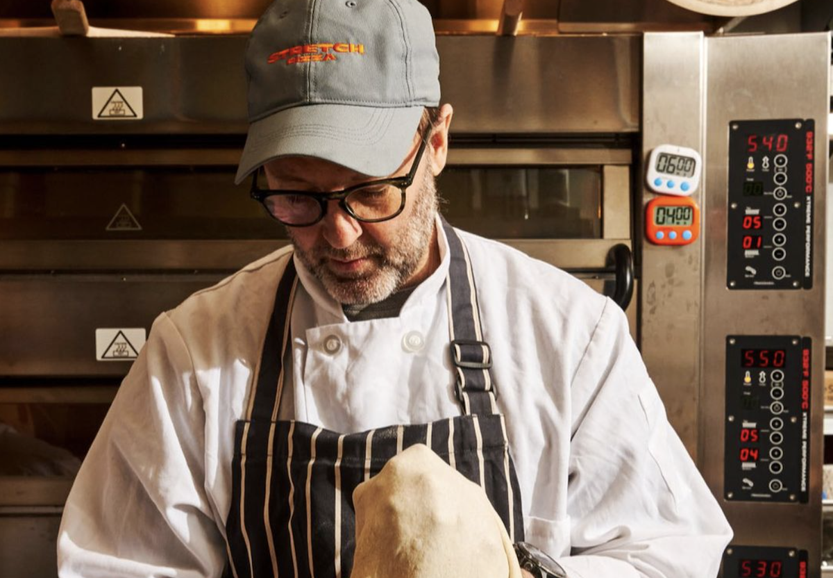 Chef Wylie Dufresne of Stretch Pizza stretches pizza dough in a kitchen, donning a chef coat and apron.