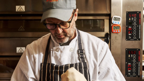 Chef Wylie Dufresne of Stretch Pizza stretches pizza dough in a kitchen, donning a chef coat and apron.