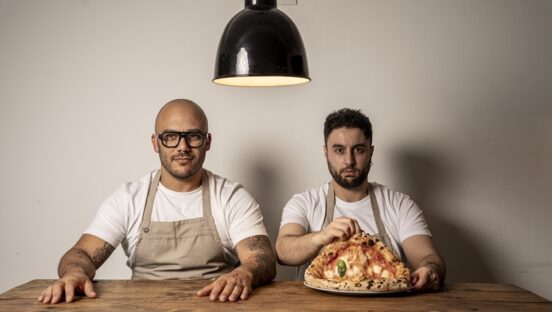 This photo shows the two owners of Sartoria Panatieri seated at a table. The bearded man on the right is displaying a large Neapolitan Margherita pizza.