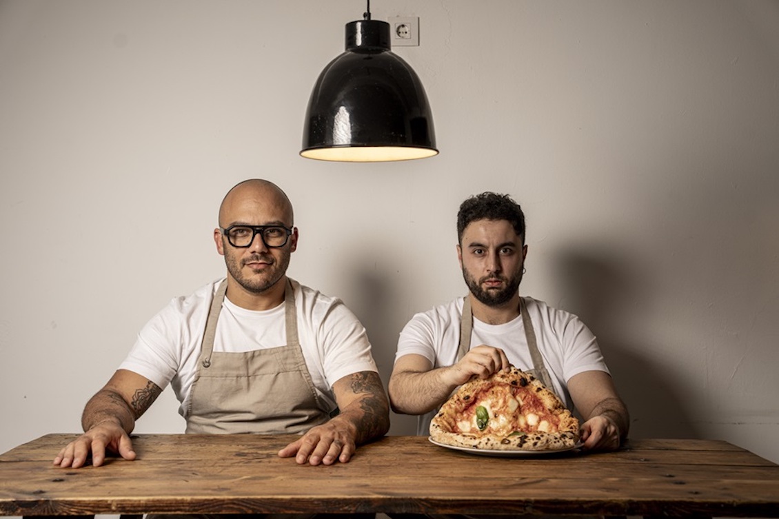 This photo shows the two owners of Sartoria Panatieri seated at a table. The bearded man on the right is displaying a large Neapolitan Margherita pizza.