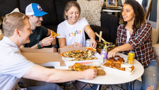 This photo shows four people gathered around a table and eating carryout pizza.