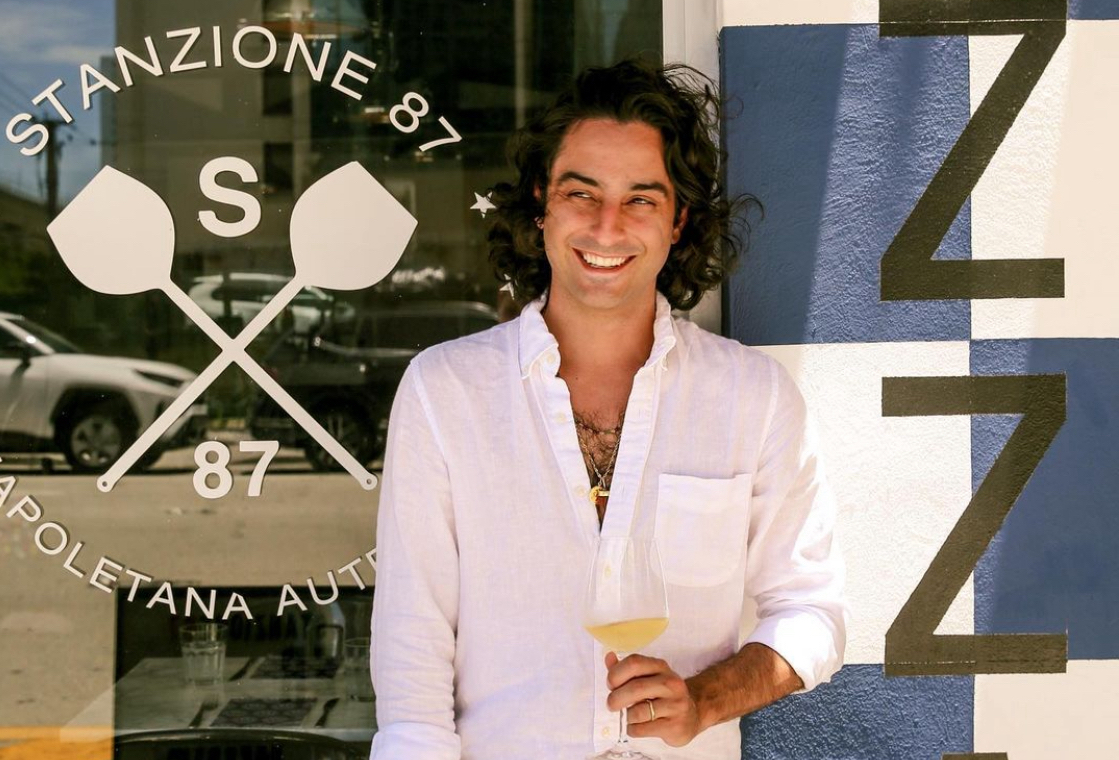 This photo shows a smiling Franco Stanzione, with long dark hair and a white shirt, standing in front of his restaurant, holding a drink in his hand.