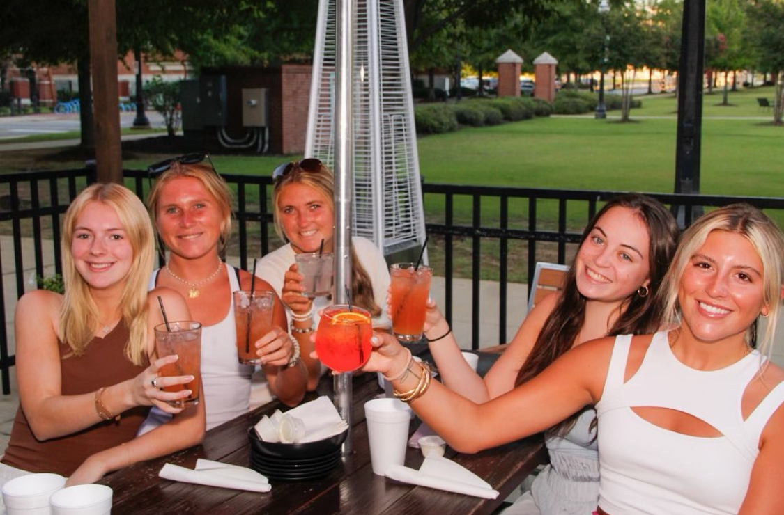 This photo shows a group of five young women sitting at Heat Pizza Bar's patio table, smiling at the camera and holding up their cocktails.