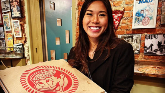 This photo shows a dark-haired woman in a dark shirt smiling and holding up a Pizza Brain pizza box inside the restaurant.