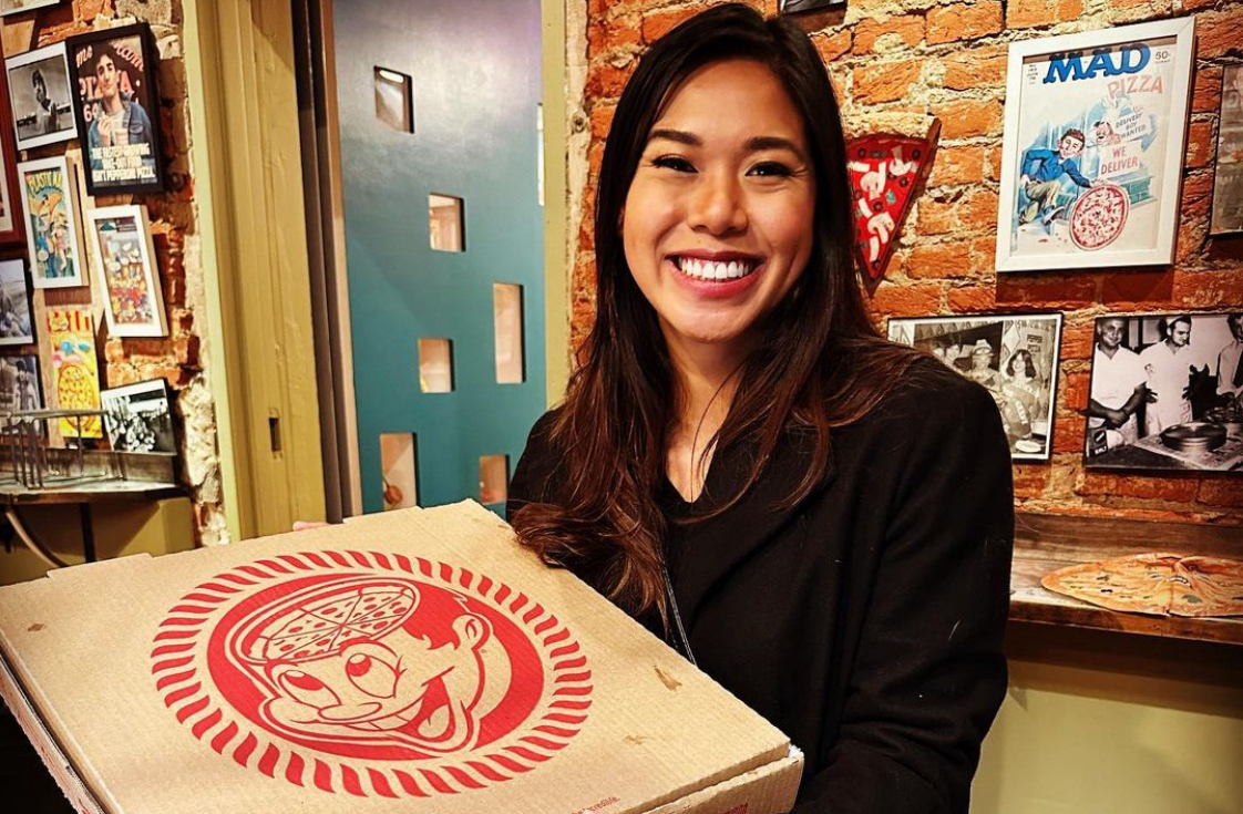This photo shows a dark-haired woman in a dark shirt smiling and holding up a Pizza Brain pizza box inside the restaurant.