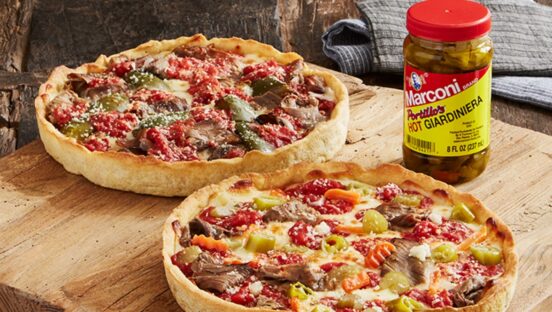 Two deep-dish pizzas sit on display next to a container of hot peppers.