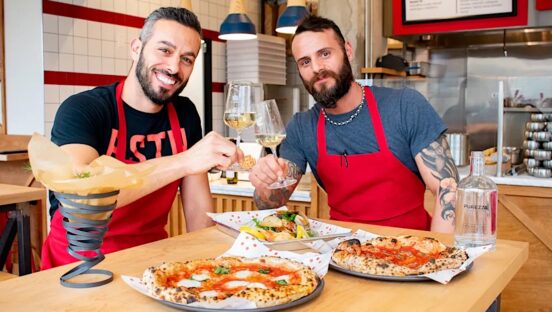 This photo shows the two owners of Stellina Pizzeria, both dark-haired and bearded, clinking their champagne glasses together with two pizzas on the table in front of them.