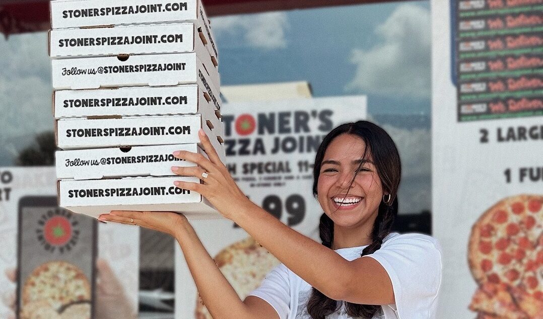 This photo shows a happily smiling woman holding up a stack of Stoner's Pizza Joint carryout boxes in front of a Stoner's restaurant.
