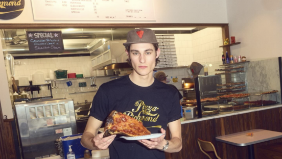 A young man stands in a pizza shop holding a slice of pepperoni pizza. He appears ready to eat it.