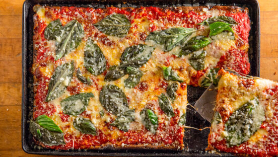 A Sicilian, square-style pizza shot from overhead. The pizza has healthy amounts of tomato sauce, cheese, and basil, and a corner square is being cut out of it.