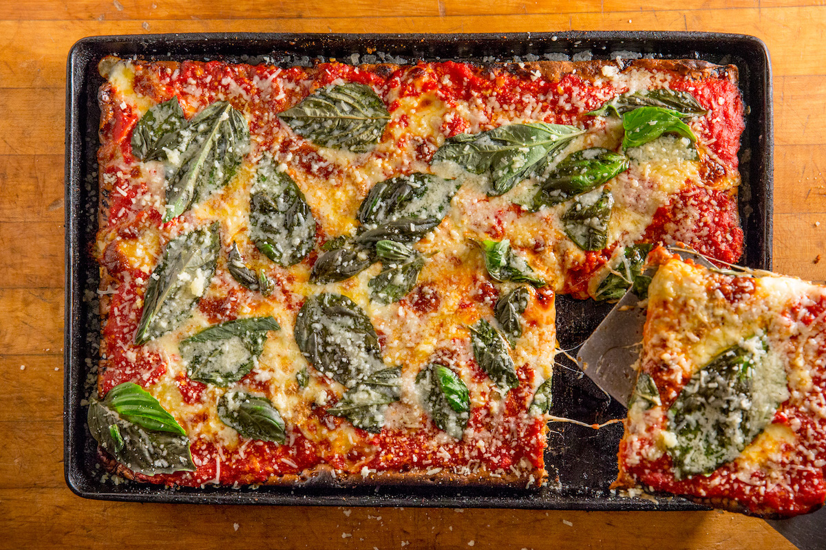 A Sicilian, square-style pizza shot from overhead. The pizza has healthy amounts of tomato sauce, cheese, and basil, and a corner square is being cut out of it.