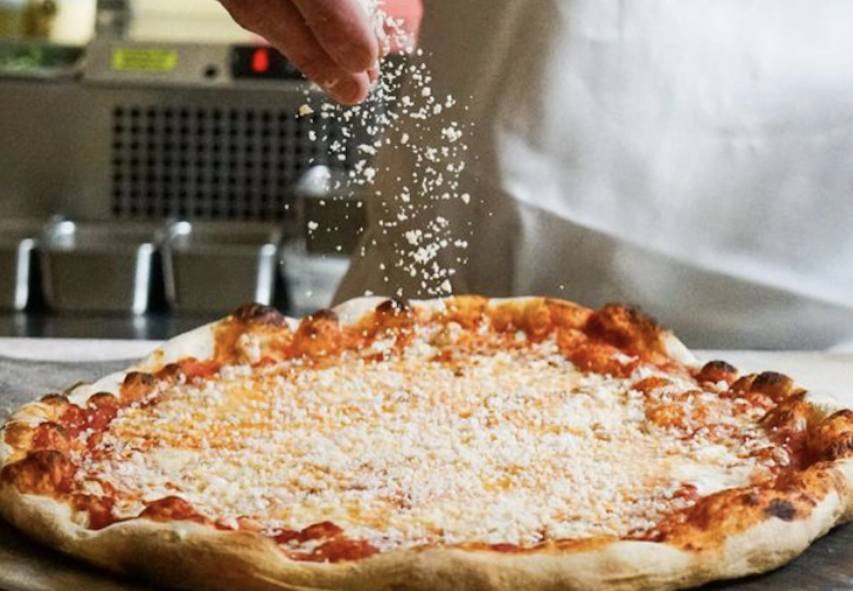 A hand sprinkles cheese across a cheese pizza.