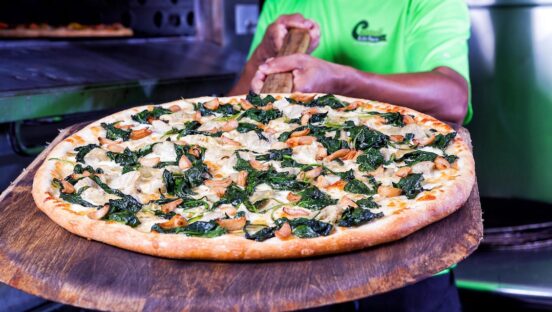 This photo shows a Black pizza maker (from the neck down) presenting a pizza on a wooden peel.
