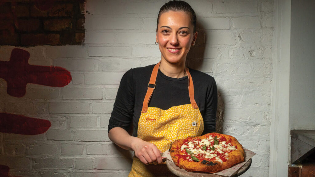This photo shows Giorgia Caporuscio smiling and holding a small peel with a Margherita pizza.