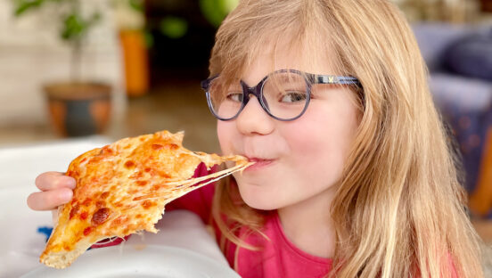 This photo shows a little girl with glasses grinning as she bites into a cheesy pizza slice.
