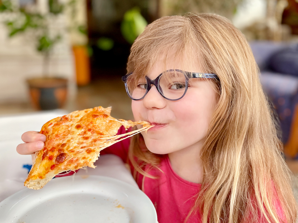 This photo shows a little girl with glasses grinning as she bites into a cheesy pizza slice.