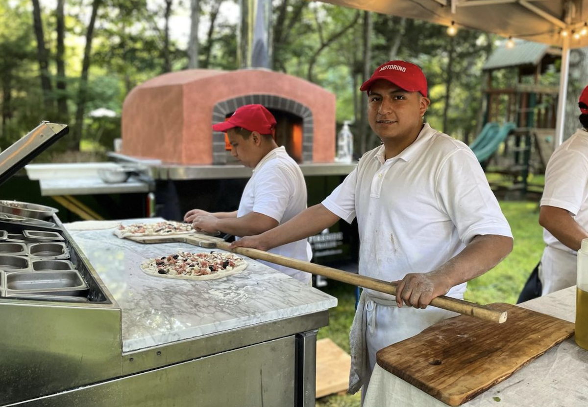 A man holds a pizza on a peel in a mobile pizza catering set up.