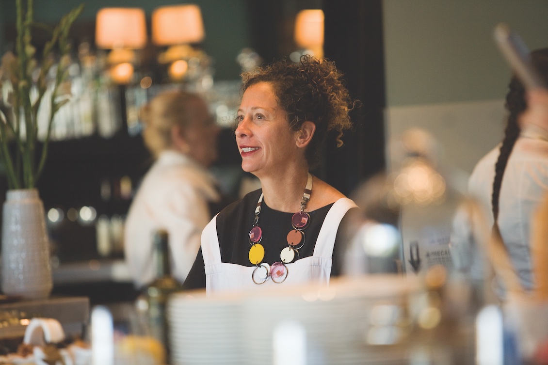 This photo shows Nancy Silverton, with her dark hair in a bun, in profile, smiling at someone.