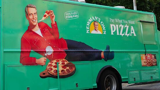 This photo shows the Newman's Own pizza truck, with an illustration of a smiling Paul Newman reclining and leaning on his elbow, eating a slice of pepperoni pizza with his free hand.