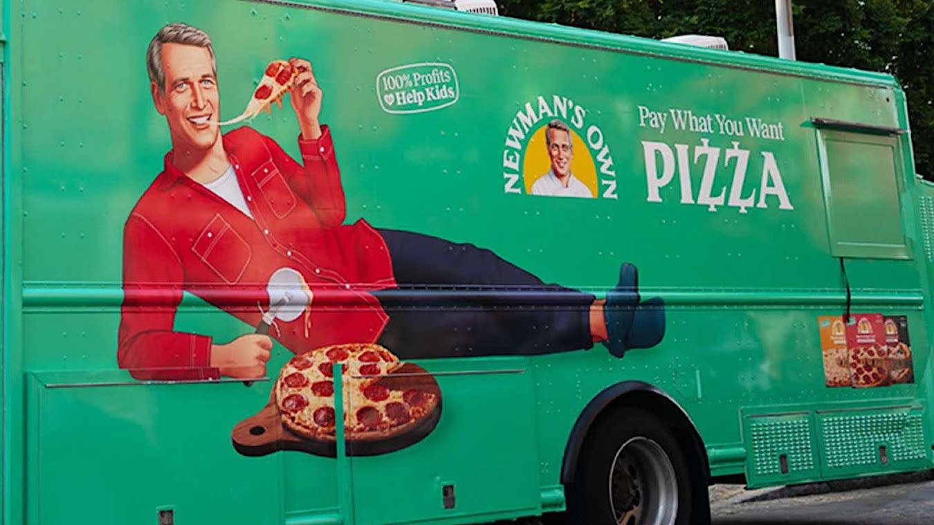 This photo shows the Newman's Own pizza truck, with an illustration of a smiling Paul Newman reclining and leaning on his elbow, eating a slice of pepperoni pizza with his free hand.