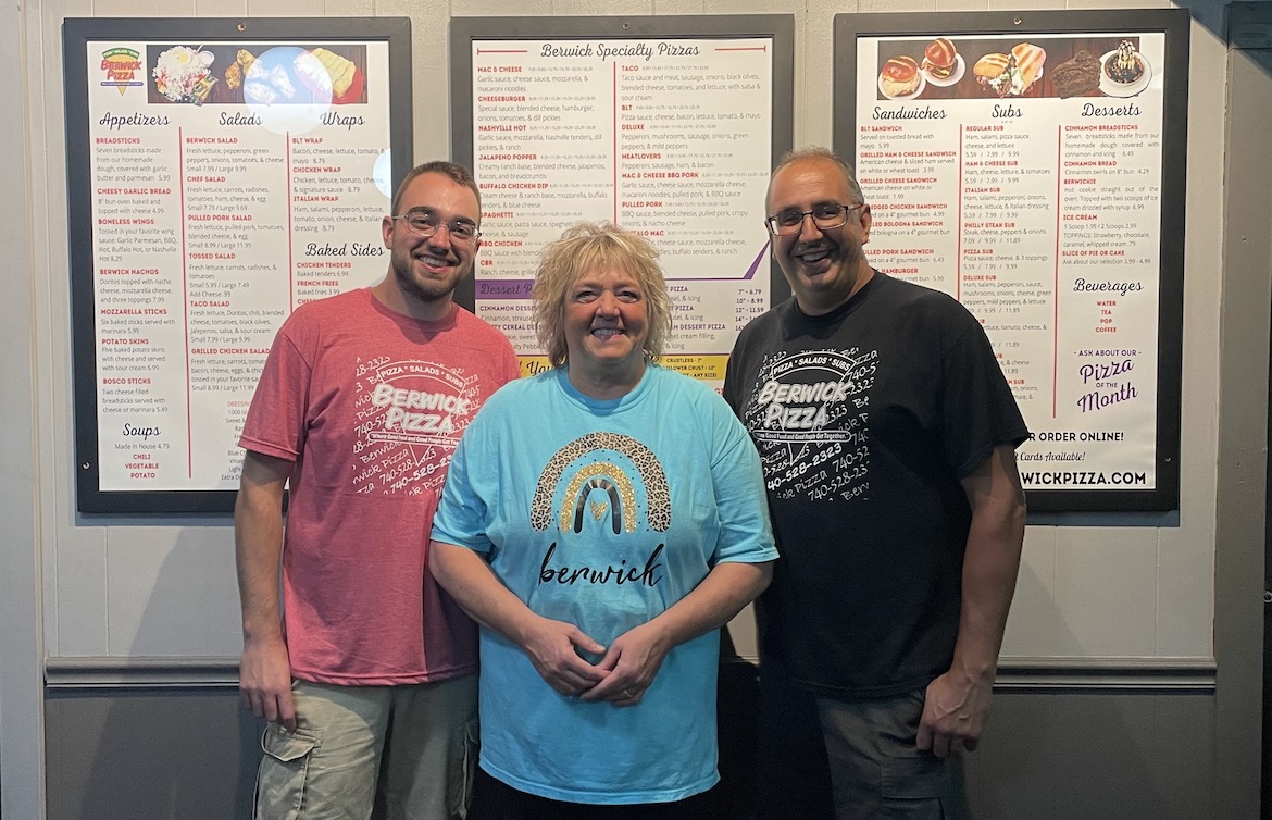 This photo shows Austin Berwick, his mother and his father, all wearing shorts and t-shirts, standing in front of Berwick Pizza's three-piece menu board in their restaurant.